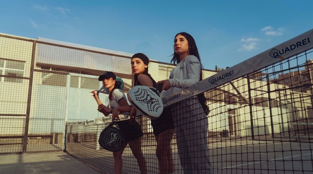 Três jogadoras de padel com uniforme do Quadrei, de raquete em punho ao lado da rede
